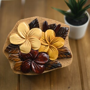 May include: A wooden box carved with three plumeria flowers. The flowers are carved in shades of brown, yellow, and red. The box is a unique and decorative piece of art.