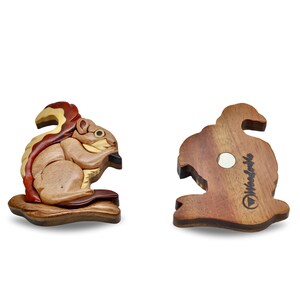 May include: Wooden squirrel figurine with a detailed, layered design. The squirrel is depicted in shades of brown, tan, and red, holding a dark object. The back of the figurine shows a magnet and the text "Woodville".