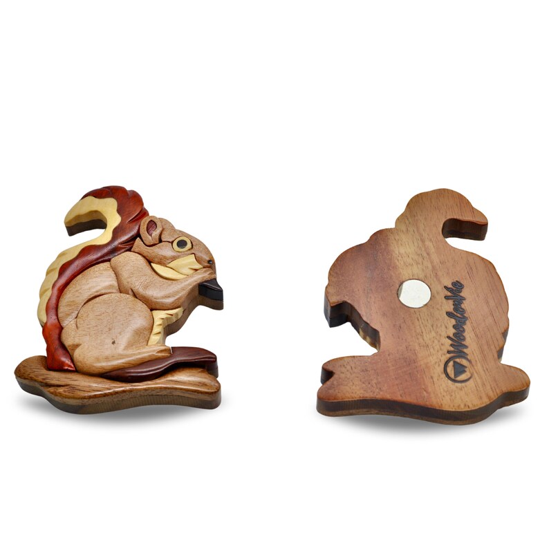 May include: Wooden squirrel figurine with a detailed, layered design. The squirrel is depicted in shades of brown, tan, and red, holding a dark object. The back of the figurine shows a magnet and the text "Woodville".