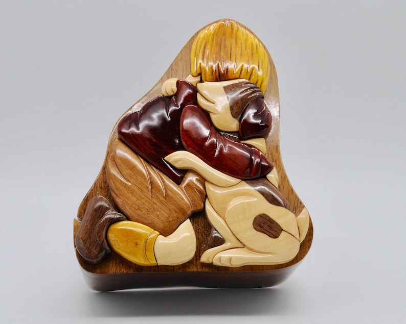 May include: A wooden puzzle box with a carved design of a child hugging a dog. The box is made of different types of wood, including light brown, dark brown, and red.