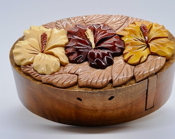 May include: A wooden puzzle box with a hinged lid, featuring three intricately carved hibiscus flowers in shades of yellow, brown, and red. The box is made of light brown wood and has a smooth, polished finish.