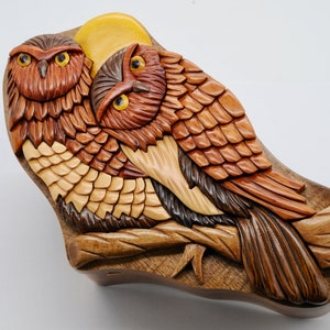 Op de afbeelding: Een houten uil puzzeldoos met twee uilen die op een tak zitten. De uilen zijn ingewikkeld gesneden met gedetailleerde veren en hebben realistische ogen. De doos is geschilderd in tinten bruin, geel en oranje.