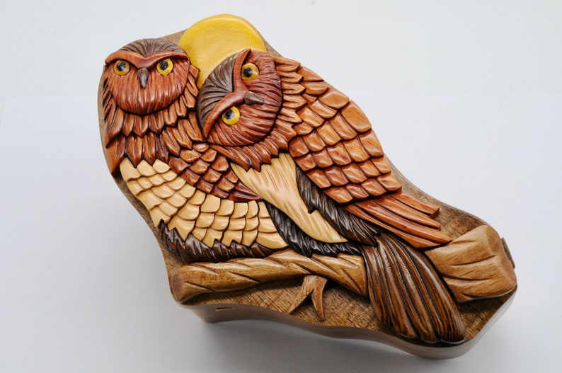 Op de afbeelding: Een houten uil puzzeldoos met twee uilen die op een tak zitten. De uilen zijn ingewikkeld gesneden met gedetailleerde veren en hebben realistische ogen. De doos is geschilderd in tinten bruin, geel en oranje.