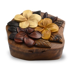 May include: A wooden puzzle box featuring a carved design of three plumeria flowers in shades of brown, yellow, and white. The box is crafted from dark wood and has a natural, rustic finish.