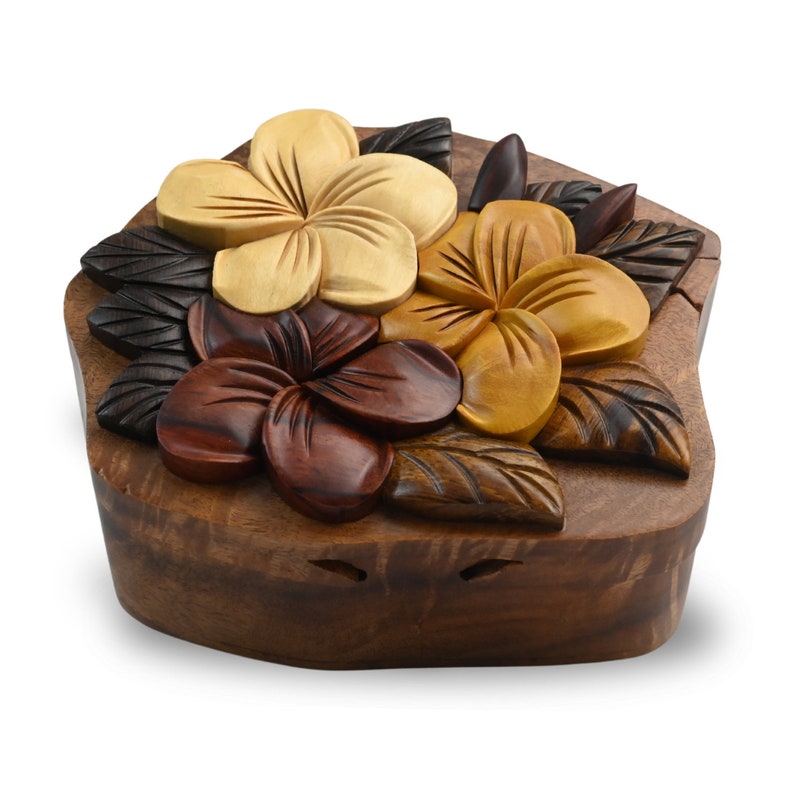 May include: A wooden puzzle box featuring a carved design of three plumeria flowers in shades of brown, yellow, and white. The box is crafted from dark wood and has a natural, rustic finish.