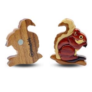 May include: Two wooden squirrel figurines. One shows the back with a magnet and the text "WoodleMe". The other is a detailed squirrel with a red body, light brown tail, and a light brown and yellow head. The squirrel is perched on a wooden branch.