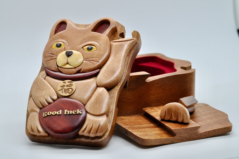 以下が含まれることがあります： 「幸運」と刻まれた赤いハートを持った茶色の猫の形をした木製パズルボックス。猫は、中国語の文字が書かれた金の首輪をしている。箱は開いており、内側は赤いベルベットで覆われている。