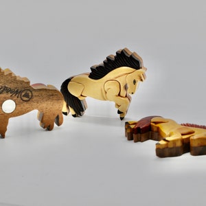 K&ouml;nnte beinhalten: Drei Holzpferde-Figuren, jede mit einer anderen Pose und Farbe. Das Pferd links ist braun mit einer schwarzen M&auml;hne und einem schwarzen Schweif, das mittlere Pferd ist hellbraun mit einer schwarzen M&auml;hne und einem schwarzen Schweif, und das Pferd rechts ist braun mit einer roten M&auml;hne und einem roten Schweif. Alle drei Pferde haben eine Holzmaserung und sind von "Woodcraft" hergestellt.