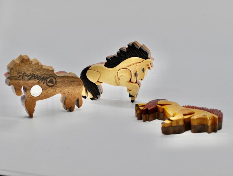 K&ouml;nnte beinhalten: Drei Holzpferde-Figuren, jede mit einer anderen Pose und Farbe. Das Pferd links ist braun mit einer schwarzen M&auml;hne und einem schwarzen Schweif, das mittlere Pferd ist hellbraun mit einer schwarzen M&auml;hne und einem schwarzen Schweif, und das Pferd rechts ist braun mit einer roten M&auml;hne und einem roten Schweif. Alle drei Pferde haben eine Holzmaserung und sind von "Woodcraft" hergestellt.