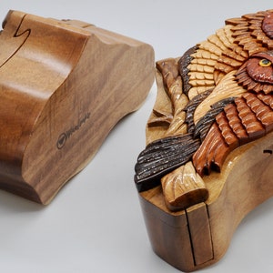 Op de afbeelding: Een houten puzzeldoos met een gesneden ontwerp van twee uilen die op een tak zitten. De doos is gemaakt van verschillende tinten bruin hout en heeft een scharnierend deksel. De doos is open en het interieur is zichtbaar.