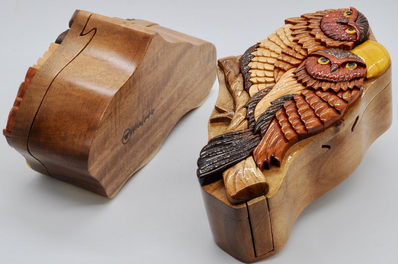 Op de afbeelding: Een houten puzzeldoos met een gesneden ontwerp van twee uilen die op een tak zitten. De doos is gemaakt van verschillende tinten bruin hout en heeft een scharnierend deksel. De doos is open en het interieur is zichtbaar.