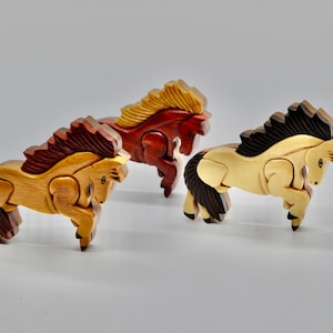 May include: Three wooden horse figurines, each carved in a different wood. The horses are in a running pose, with their heads turned to the side. The horses are painted in natural wood tones, with the mane and tail of each horse painted in a different color. The horses are arranged in a row, with the brown horse in the front, the red horse in the middle, and the tan horse in the back.