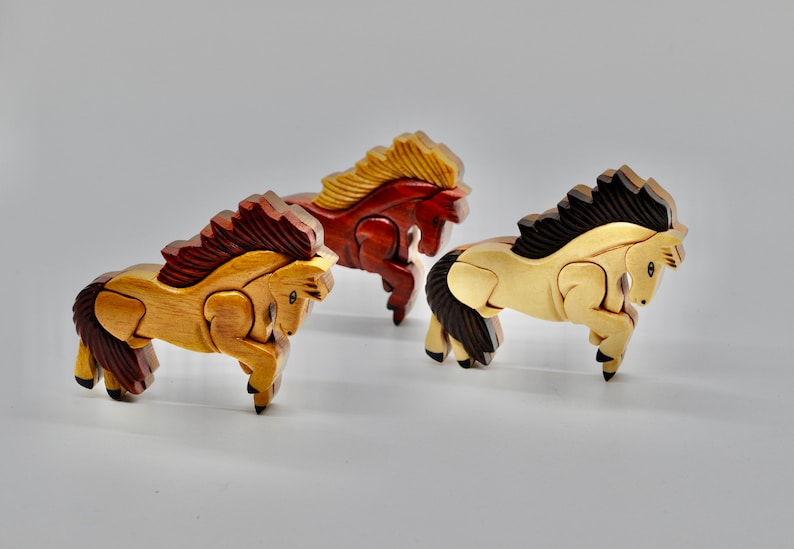 K&ouml;nnte beinhalten: Drei Holzpferde-Figuren, jede aus einem anderen Holz geschnitzt. Die Pferde sind in einer Laufhaltung, mit dem Kopf zur Seite gedreht. Die Pferde sind in nat&uuml;rlichen Holzt&ouml;nen lackiert, wobei die M&auml;hne und der Schweif jedes Pferdes in einer anderen Farbe lackiert sind. Die Pferde sind in einer Reihe angeordnet, mit dem braunen Pferd vorne, dem roten Pferd in der Mitte und dem hellbraunen Pferd hinten.