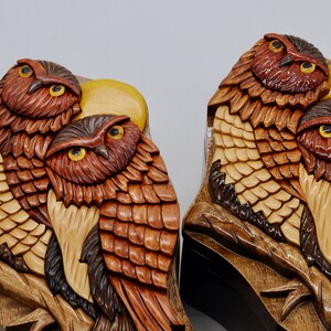 Op de afbeelding: Twee ingewikkeld gesneden houten uil puzzeldozen met een gedetailleerd ontwerp van veren en een maan. De uilen zijn bruin, rood en geel.