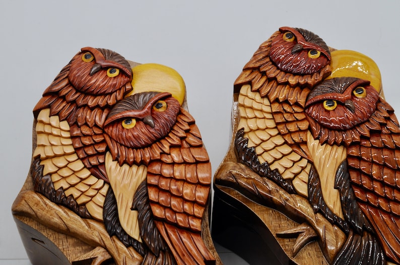 Op de afbeelding: Twee ingewikkeld gesneden houten uil puzzeldozen met een gedetailleerd ontwerp van veren en een maan. De uilen zijn bruin, rood en geel.