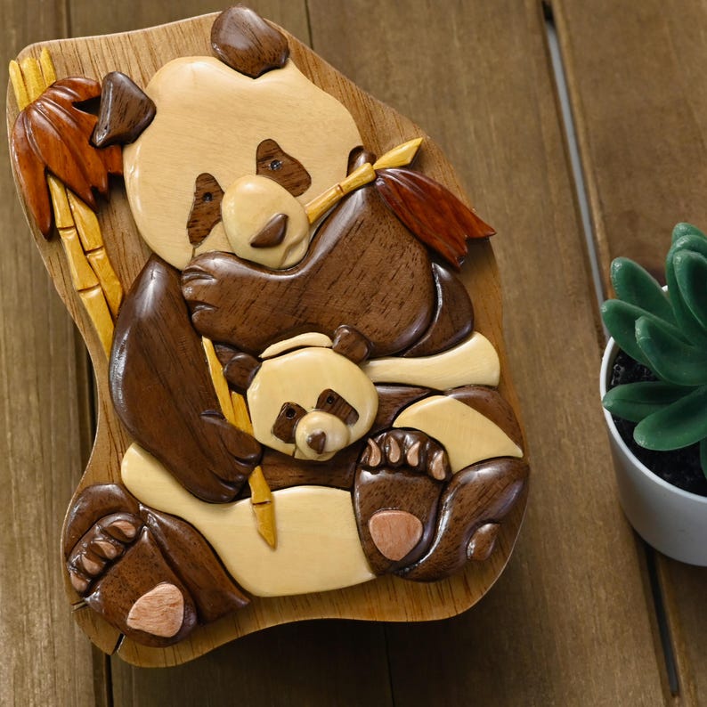 Puede incluir: Una caja de rompecabezas de madera con forma de oso panda sosteniendo un palo de bamb&uacute;. El panda tiene un patr&oacute;n de pelaje blanco y negro y est&aacute; sosteniendo una cr&iacute;a de panda m&aacute;s peque&ntilde;a. La caja est&aacute; tallada de forma intrincada y tiene un acabado de madera natural.