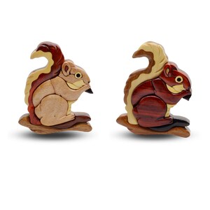 May include: Two wooden squirrel figurines, each perched on a small branch. One squirrel is primarily light brown with a dark red tail, while the other is dark red with a light brown tail. Both have black noses and are made of layered wood.