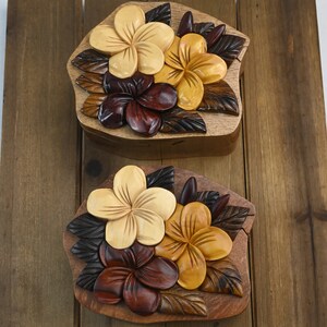 May include: Two wooden puzzle boxes with intricate carvings of flowers. The boxes are made of different shades of brown wood and feature a variety of flowers, including yellow, orange, and red.