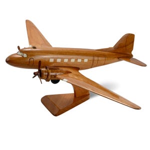 K&ouml;nnte beinhalten: Ein Holzmodell eines Vintage-Flugzeugs mit brauner Oberfl&auml;che und wei&szlig;en Akzenten. Das Modell steht auf einem Holzsockel.