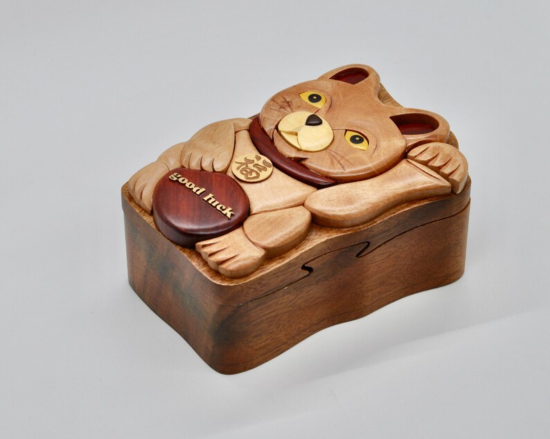 以下が含まれることがあります： 猫の形をした木製のパズルボックス。赤い金色のコインが入っています。クルミ材で作られており、猫の顔と足が細かく彫刻されています。箱には「幸運」と刻まれています。