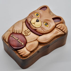 以下が含まれることがあります： 招き猫の形をした木製パズルボックス。前脚を上げており、首輪には金貨が付いています。箱は明るい木と暗い木で作られており、前面には「幸運」と書かれた赤い木製の円があります。