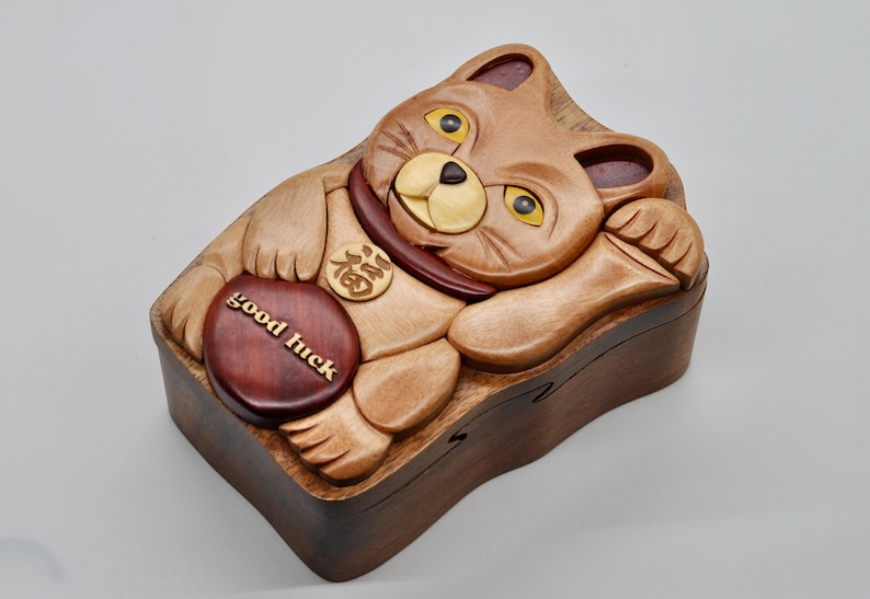 以下が含まれることがあります： 招き猫の形をした木製パズルボックス。前脚を上げており、首輪には金貨が付いています。箱は明るい木と暗い木で作られており、前面には「幸運」と書かれた赤い木製の円があります。