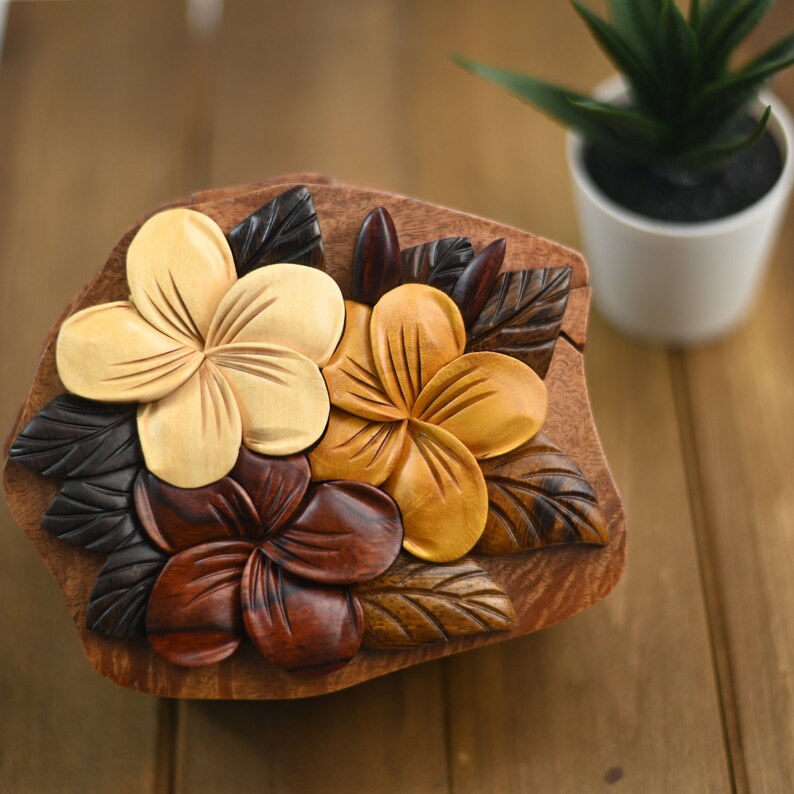 May include: A wooden puzzle box featuring a carved design of three plumeria flowers in shades of brown, yellow, and white. The box is shaped like a leaf and has a natural wood finish.