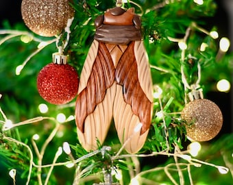 May include: A wooden ornament shaped like a cicada with brown, beige, and reddish-brown wood tones. The ornament is hanging on a Christmas tree with green needles and white lights.