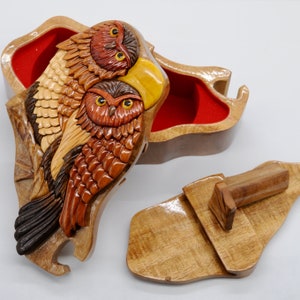 Op de afbeelding: Een houten uil puzzeldoos met een rood fluwelen interieur. De doos is ingewikkeld gesneden met twee uilen, waarvan er &eacute;&eacute;n een gele snavel heeft, en de deksel is open om het interieur te laten zien.