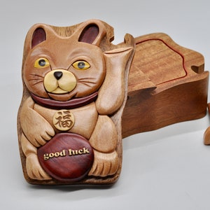 以下が含まれることがあります： 茶色と黄色の招き猫の形をした木製のパズルボックス。赤い首輪と胸に「福」の漢字が書かれた金貨が付いています。箱は開いていて、内側が見えます。蓋は別になっています。箱の前面には「幸運」の文字が刻まれています。