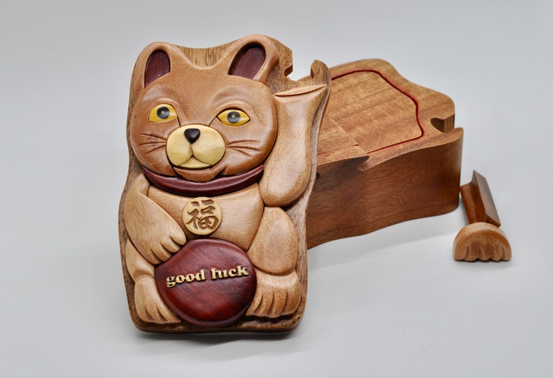 以下が含まれることがあります： 茶色と黄色の招き猫の形をした木製のパズルボックス。赤い首輪と胸に「福」の漢字が書かれた金貨が付いています。箱は開いていて、内側が見えます。蓋は別になっています。箱の前面には「幸運」の文字が刻まれています。