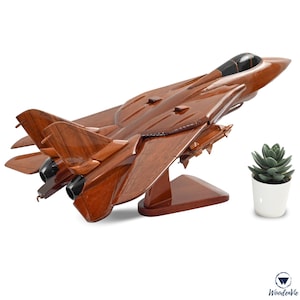 May include: A detailed wooden model of a fighter jet, finished in a dark brown stain. The model has intricate wing and engine details, and a black cockpit. It is displayed on a matching wooden stand. A small succulent in a white pot is to the right.