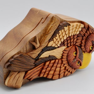 Op de afbeelding: Een houten uil puzzeldoos met ingewikkelde houtsnijwerk van drie uilen met gele ogen. De doos is gemaakt van verschillende tinten bruin hout en heeft een gele accent.
