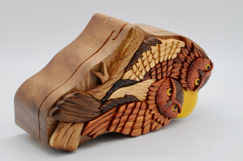 Op de afbeelding: Een houten uil puzzeldoos met ingewikkelde houtsnijwerk van drie uilen met gele ogen. De doos is gemaakt van verschillende tinten bruin hout en heeft een gele accent.