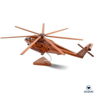 May include: A finely crafted wooden helicopter model, with a rich brown body and lighter brown rotor blades. The model is displayed on a wooden stand with a metal support. The design highlights the aircraft's detailed features.