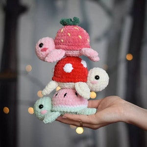 Può includere: Una pila di quattro giocattoli di tartaruga all'uncinetto. La tartaruga in cima è rosa con una fragola sul guscio. La tartaruga successiva è rossa con pois bianchi. La terza tartaruga è rosa e bianca con un design a cocomero. La tartaruga in basso è verde.