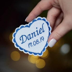 Könnte beinhalten: Ein weißer herzförmiger Patch mit einem blauen gewellten Rand. Der Patch ist mit dem Namen "Daniel" und dem Datum "17.08.19" bestickt.