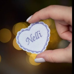 Könnte beinhalten: Ein weißer herzförmiger Patch mit einem lila bestickten Rand und dem Namen "Nelli" in lila Garn.