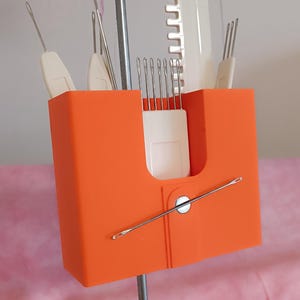 Può includere: Un porta aghi da maglieria in plastica arancione con vari strumenti per lavorare a maglia. Il supporto contiene diversi aghi con manico bianco, uno strumento bianco a forma di pettine e un ago lungo. Il supporto è montato su un'asta di metallo.