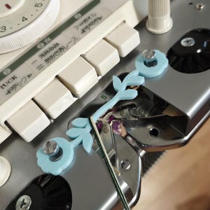 May include: Close-up of a vintage knitting machine with a light blue floral accessory. The machine has white keys, a dial, and Japanese text. The accessory is attached with a silver screw.