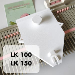 May include: A white knitting machine accessory with a textured surface and a circular knob. The accessory is positioned over a gray and pink knitting machine. The image also includes a card with the text "my creative land handmade studio" and the text "LK 100 LK 150".