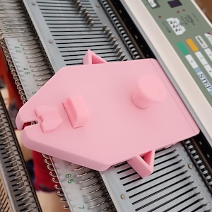Puede incluir: Una herramienta rosa claro para máquina de tejer con un diseño plano y angular. Cuenta con un botón central y varios elementos salientes, que descansan sobre una base de máquina de tejer gris y plateada. La herramienta está diseñada para ayudar a tejer.