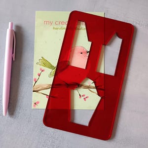 Puede incluir: Una plantilla de letras de acrílico rojo con un recorte de la letra "A" se coloca sobre una tarjeta de felicitación con una ilustración de un pájaro. Un bolígrafo rosa está a la izquierda. La tarjeta dice "my creative tone handmade studio".