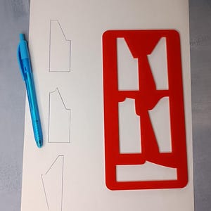 May include: A sheet of paper with three pencil-drawn garment pattern outlines, a red plastic pattern stencil, and a blue pen. The stencil has cut-out shapes matching the drawn outlines. The pen is lying on the paper.
