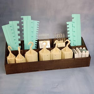 Könnte beinhalten: Ein dunkler Holz-Organizer mit mehreren Fächern, gefüllt mit verschiedenen Bastelutensilien. Der Organizer enthält hellgrüne Kunststoffwerkzeuge, cremefarbene Haken und Nadeln. Die Artikel sind ordentlich angeordnet, was auf Organisation und Handwerk hindeutet.