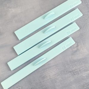 Könnte beinhalten: Vier hellblaue, rechteckige Werkzeuge mit abgerundeten Griffen. Jedes Werkzeug ist mit einem Maß in Millimetern beschriftet: 18 mm, 20 mm, 22 mm und 24 mm. Die Werkzeuge sind diagonal auf einer strukturierten, grauen Oberfläche angeordnet.