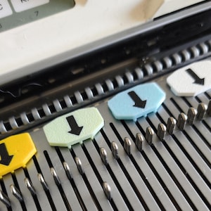 Könnte beinhalten: Ein Nadelbett einer Strickmaschine mit einer Reihe metallischer Nadeln und mehreren farbigen Kunststoffmarkern. Die Marker sind gelb, hellgrün und hellblau und zeigen jeweils einen schwarzen Pfeil, der nach unten gerichtet ist.