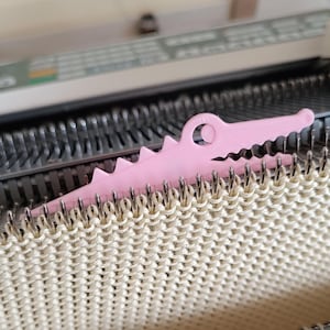 May include: A light pink, crocodile-shaped knitting tool rests on a cream-coloured knitted fabric. The tool has a circular opening and a jagged spine. The background shows a knitting machine with metal needles.