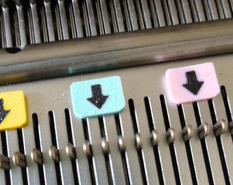 May include: A metallic knitting machine needle bed featuring a series of vertical slots and metal needles. Three small, rectangular plastic markers in yellow, light blue, and light pink are attached to the needles, each displaying a black arrow.