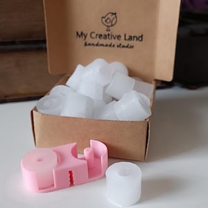 Peut inclure: Une boîte en carton marron contenant des objets cylindriques blancs. Un outil rose et un cylindre blanc sont devant la boîte. Le texte "My Creative Land handmade studio" et un logo d'oiseau sont visibles.